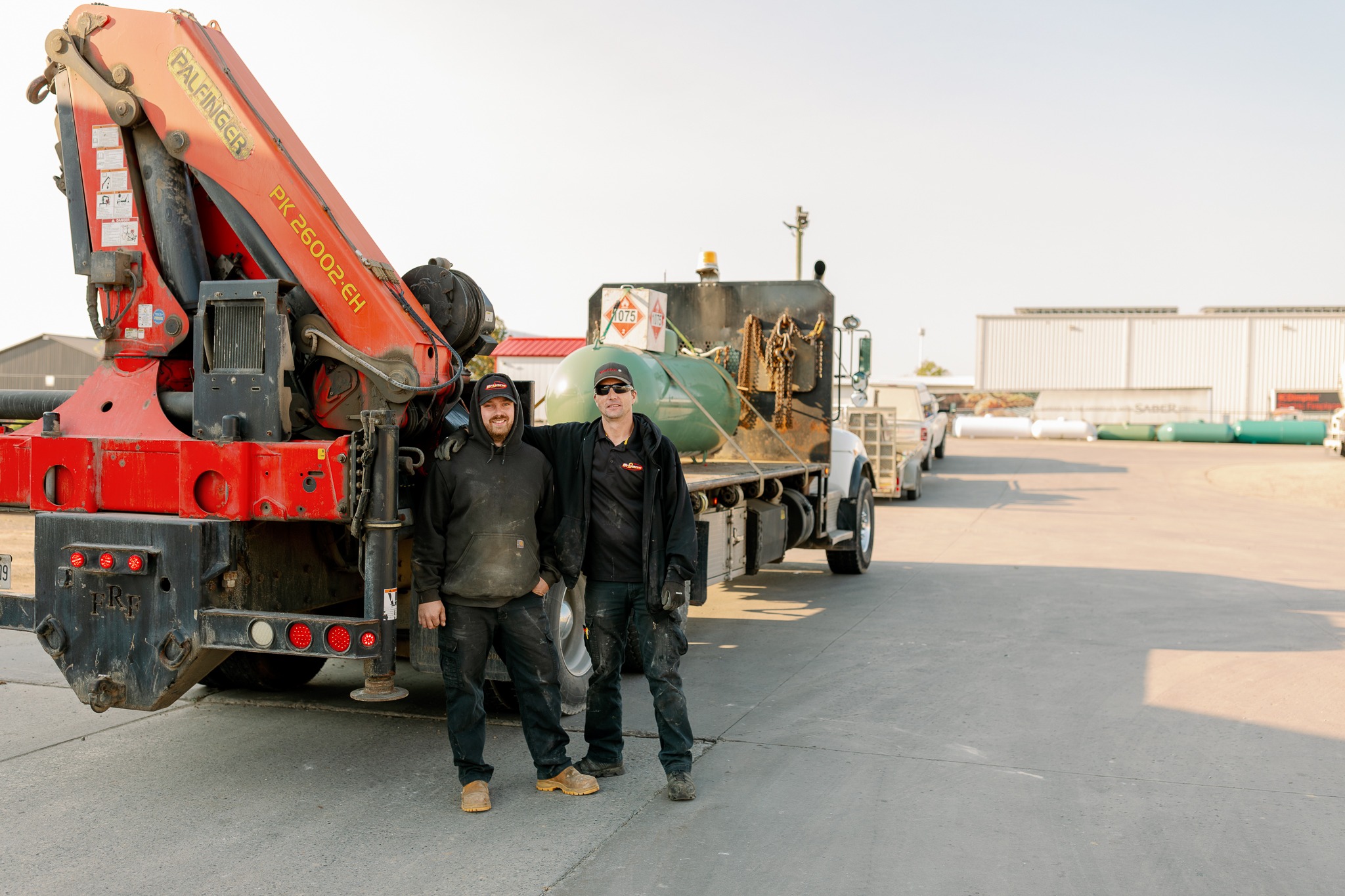 Two people stand next to a truck with construction equipment in an industrial area, wearing work attire and smiling.