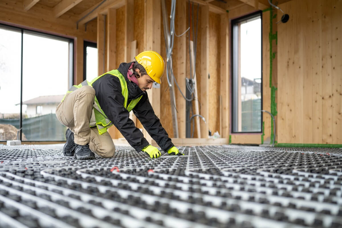 A person in a hard hat and vest works on a construction project, kneeling to install underfloor systems in a wooden building.