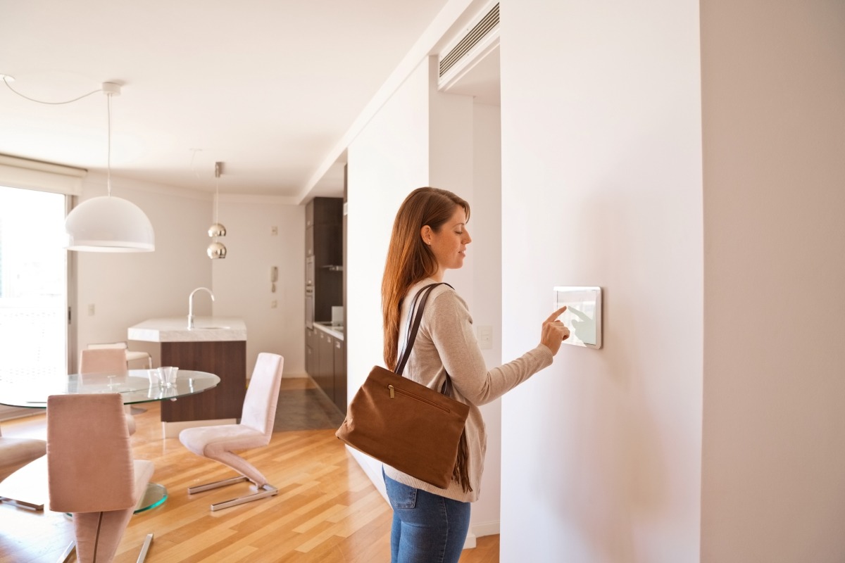 A person with a brown bag interacts with a smart home panel in a modern, well-lit kitchen featuring wooden floors and sleek furniture.