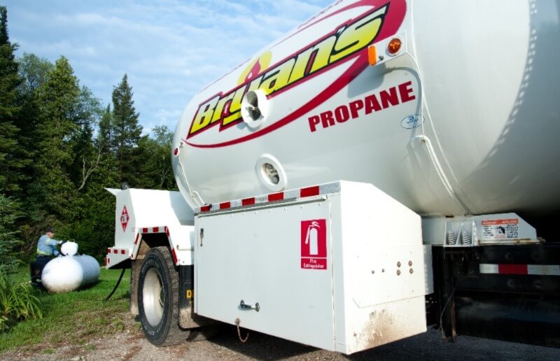A propane delivery truck with a large tank and branded logo is parked outdoors; a person works near a small white tank.