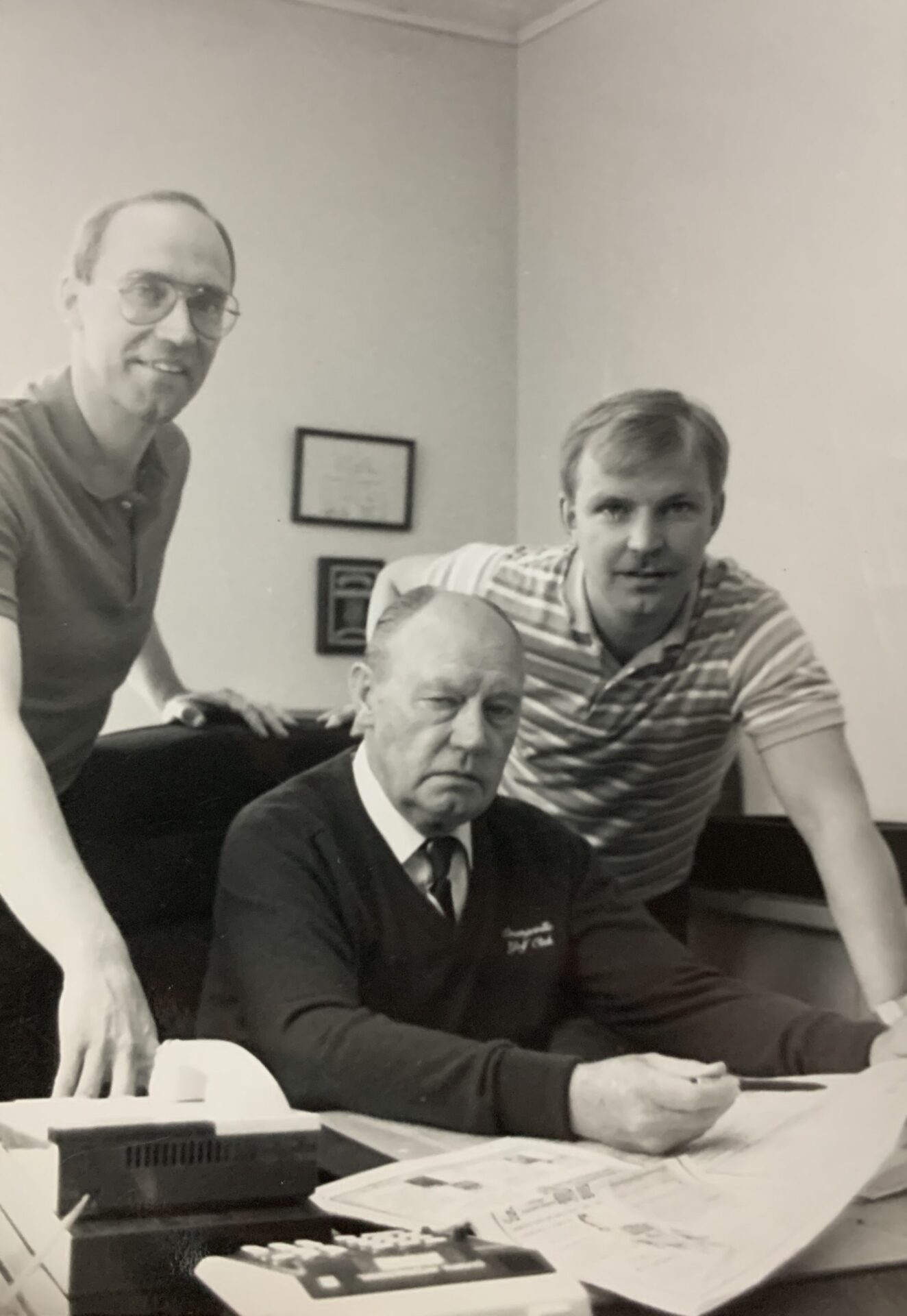 Three people gather around a desk in an office setting, with one person seated, holding a newspaper, and paperwork visible.