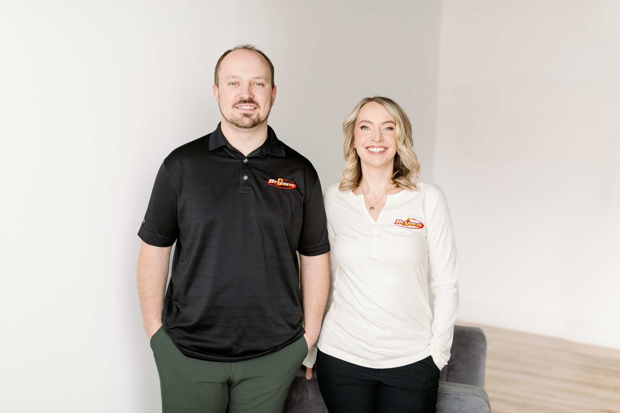 Two people smiling, wearing Besam-branded shirts, standing in a minimalist, light-toned room with a wooden floor and a gray chair.