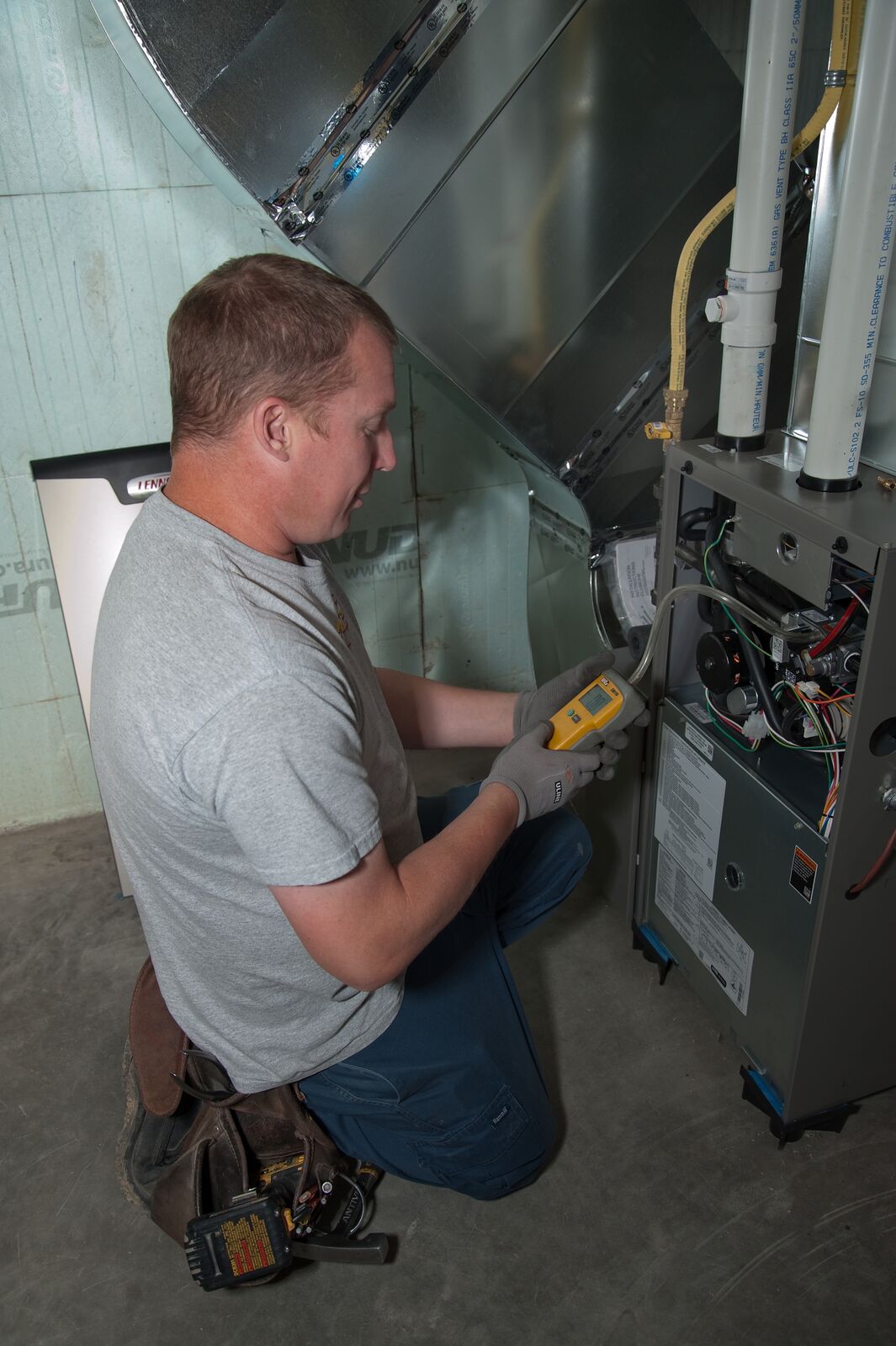 A person in a gray shirt uses a digital device to inspect a furnace unit in a basement setting with metal ductwork.