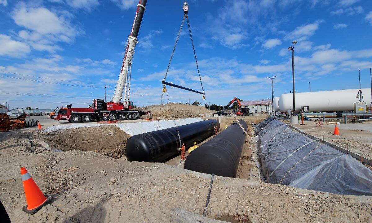 A construction site with large tanks being installed by a crane. Multiple workers and construction vehicles are present under a blue sky.