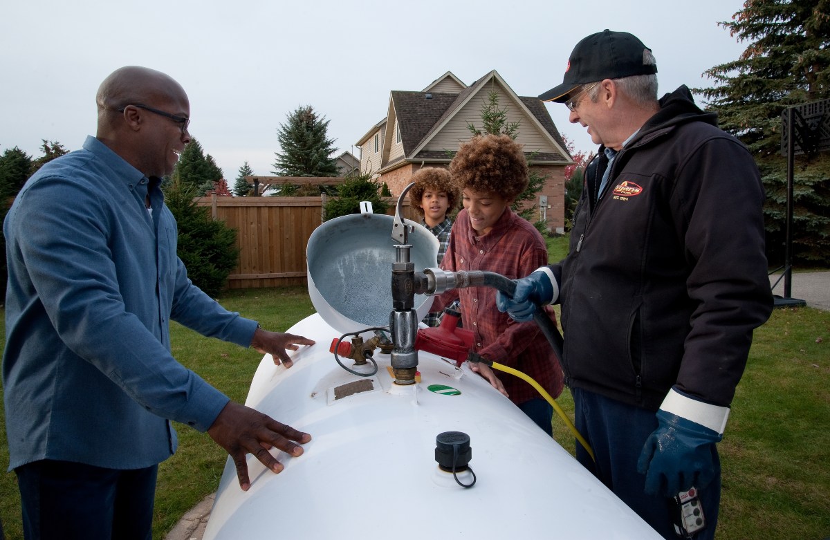 People gather around a large white tank in a suburban backyard, engaging with equipment, surrounded by trees and houses.
