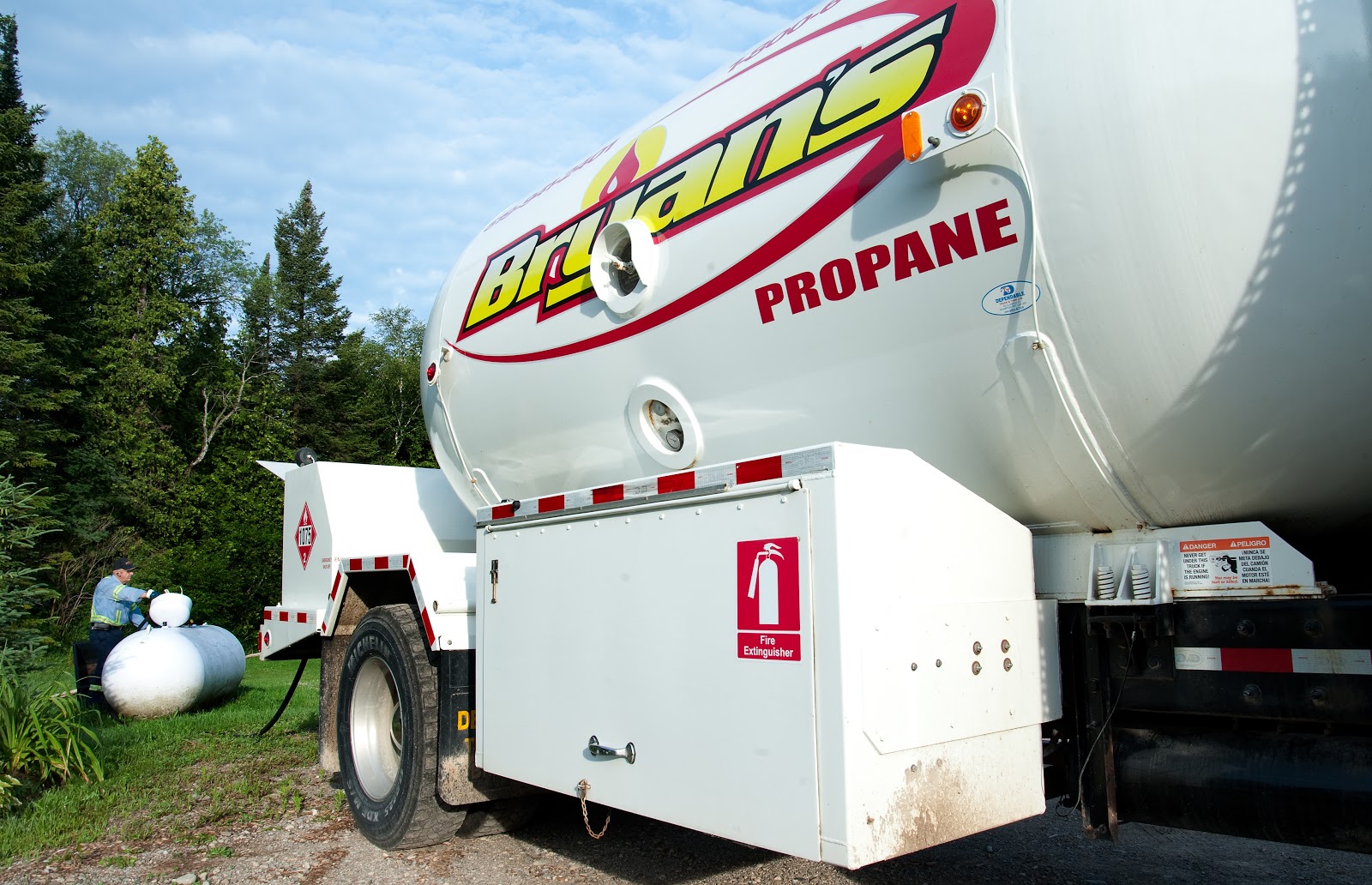 A large propane delivery truck parked near a wooded area, with a person in the background handling a propane tank.