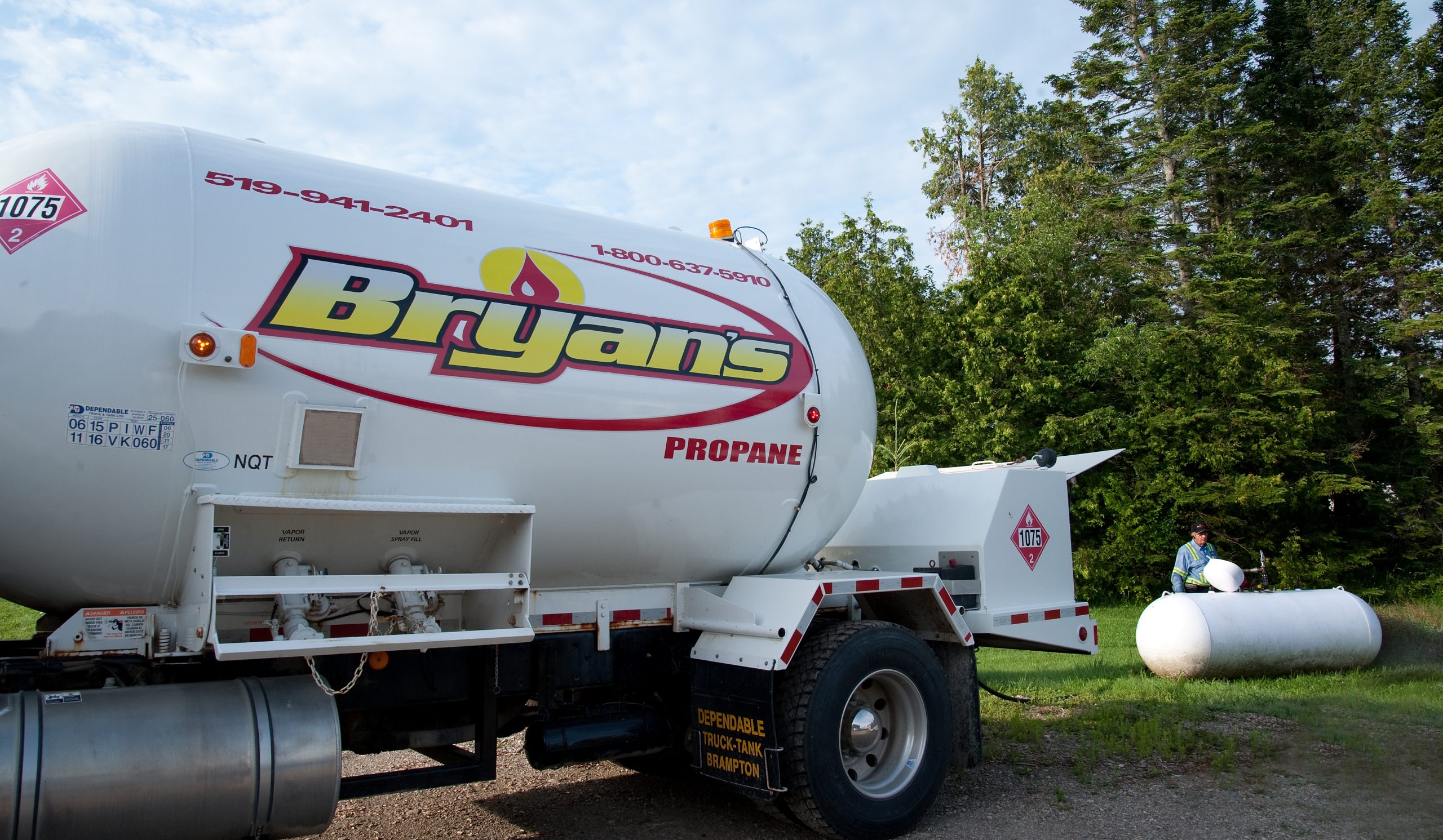 A propane truck labeled "Bryan's" is parked near a green area. A person in a safety vest interacts with equipment beside it.