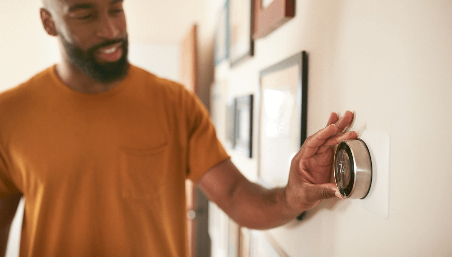A person adjusts a smart thermostat on a wall, surrounded by framed pictures, displaying a temperature of 74 degrees.