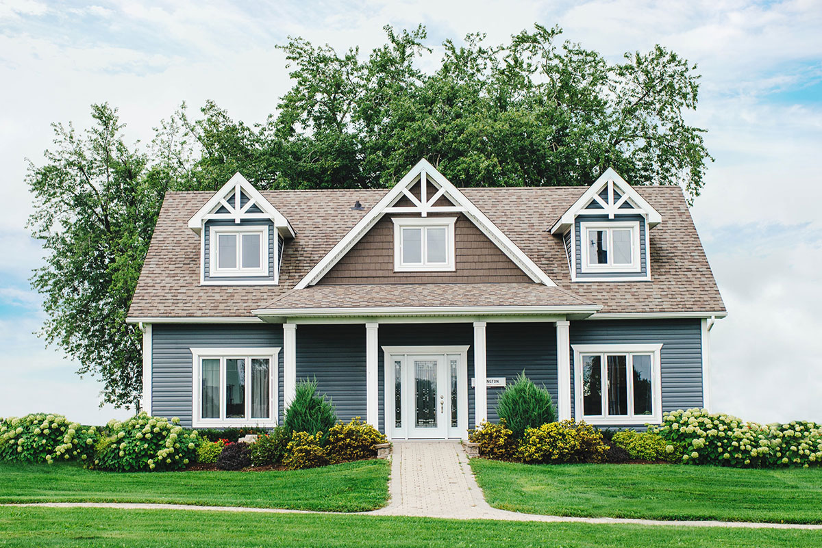 A charming blue house with white trim, surrounded by lush greenery and flowers, under a partly cloudy sky.