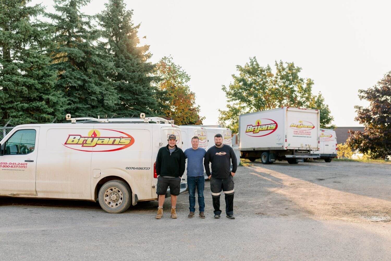 Three people stand in front of Bryans service vehicles, surrounded by trees and parked trucks under a clear sky.