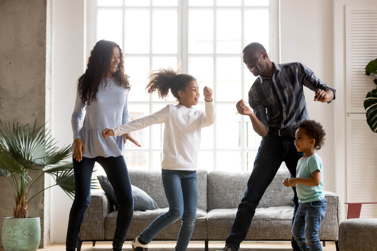 Four people dancing joyfully in a bright living room, with large windows and comfortable furniture in the background.