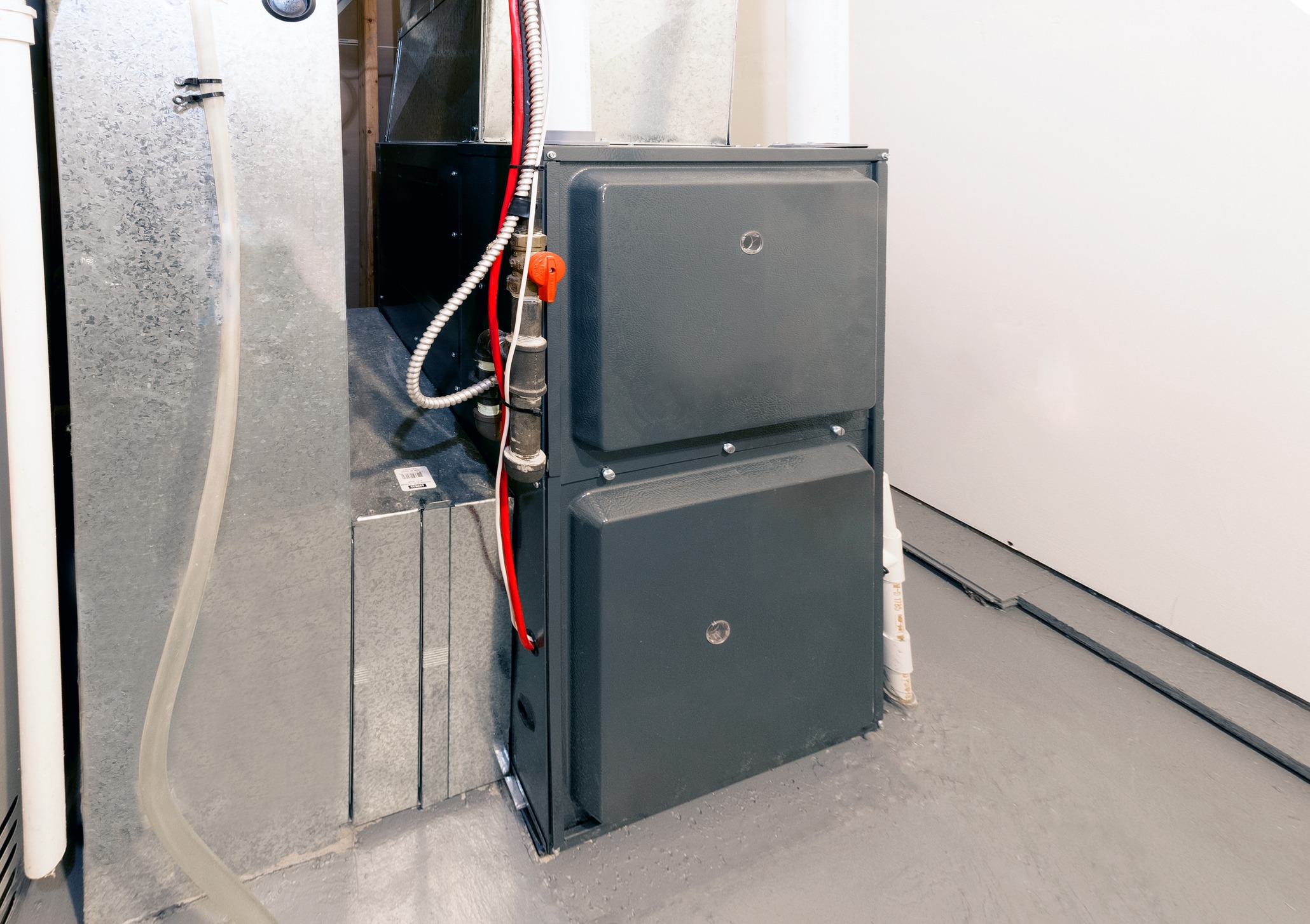 A large gray HVAC unit is installed in a room with metal ducts, red wires, and a white pipe.