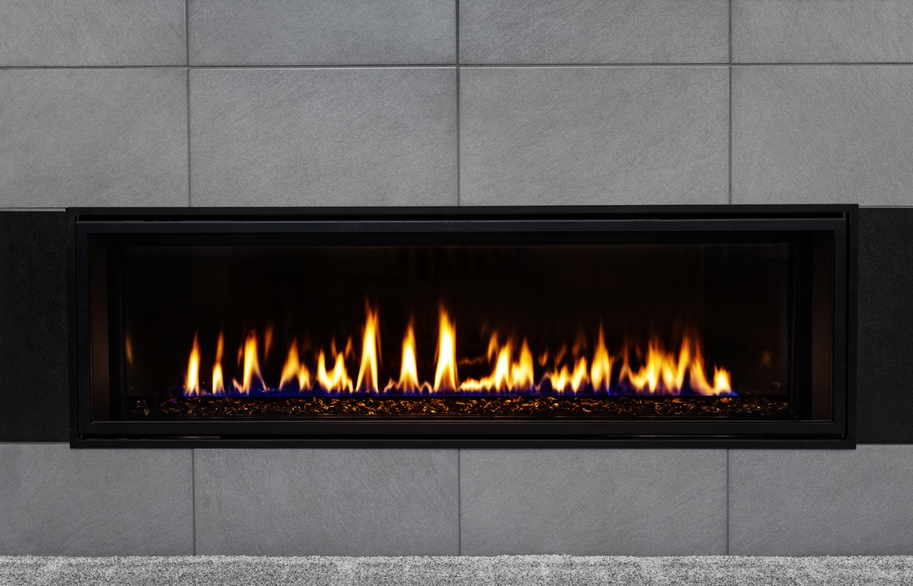 A modern linear gas fireplace set against a gray tiled wall, radiating warmth with its vibrant orange and blue flames.