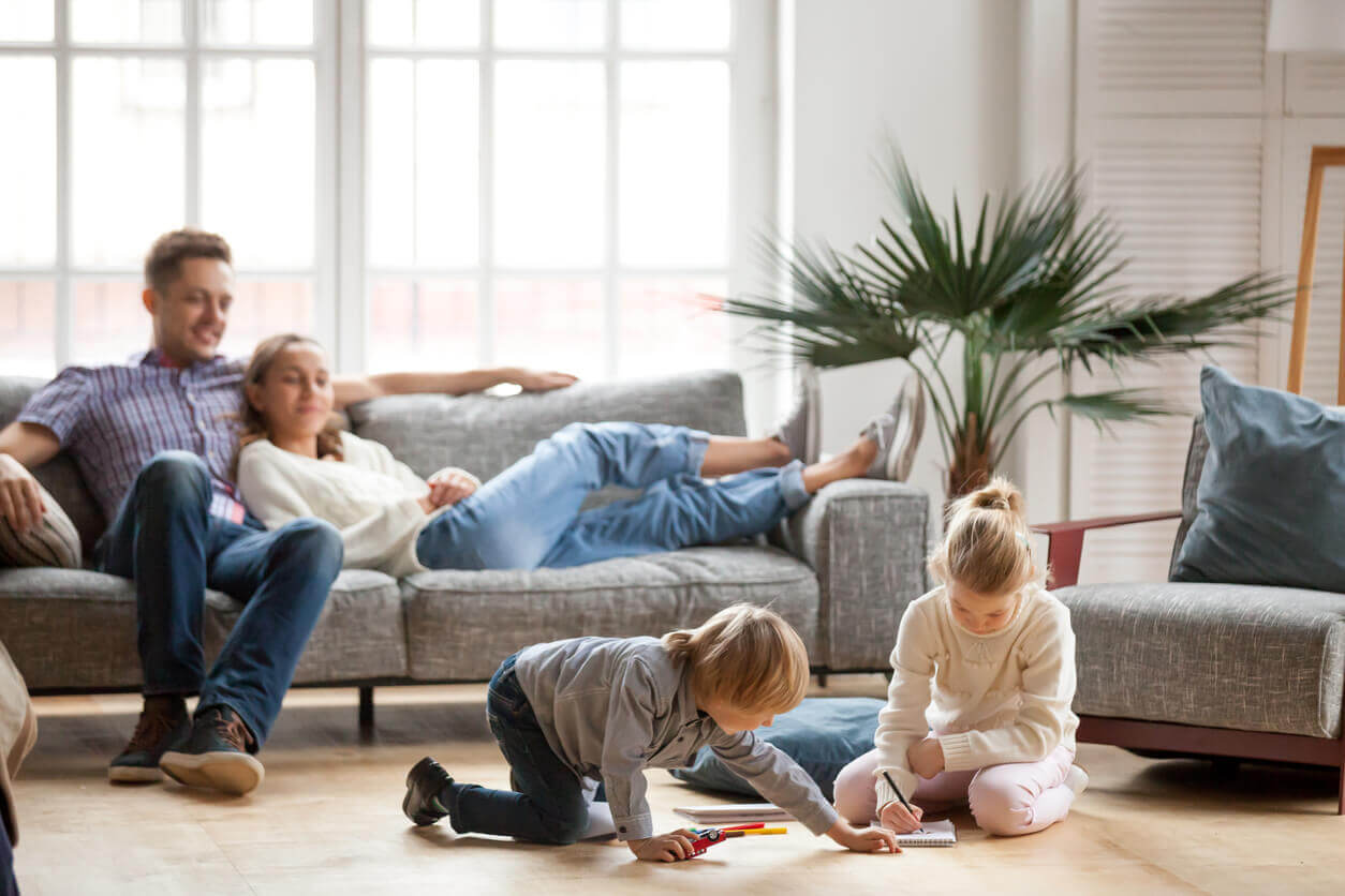 Two children draw on the floor as two people relax on a couch in a cozy, bright room with a large window.