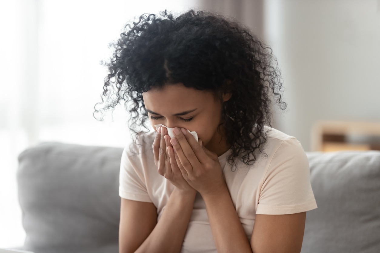 A person with curly hair sits on a sofa, using a tissue while appearing to sneeze or have a cold.