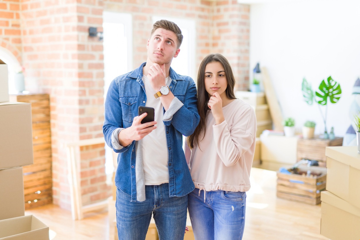 Two people stand thoughtfully in a bright room surrounded by moving boxes, one holding a phone. Plants decorate the space.