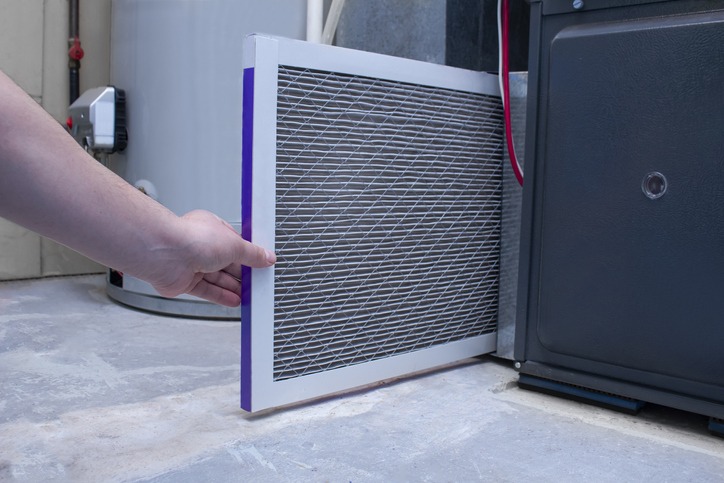 A person is changing an air filter in an HVAC system. The scene is indoors, with a visible water heater in the background.