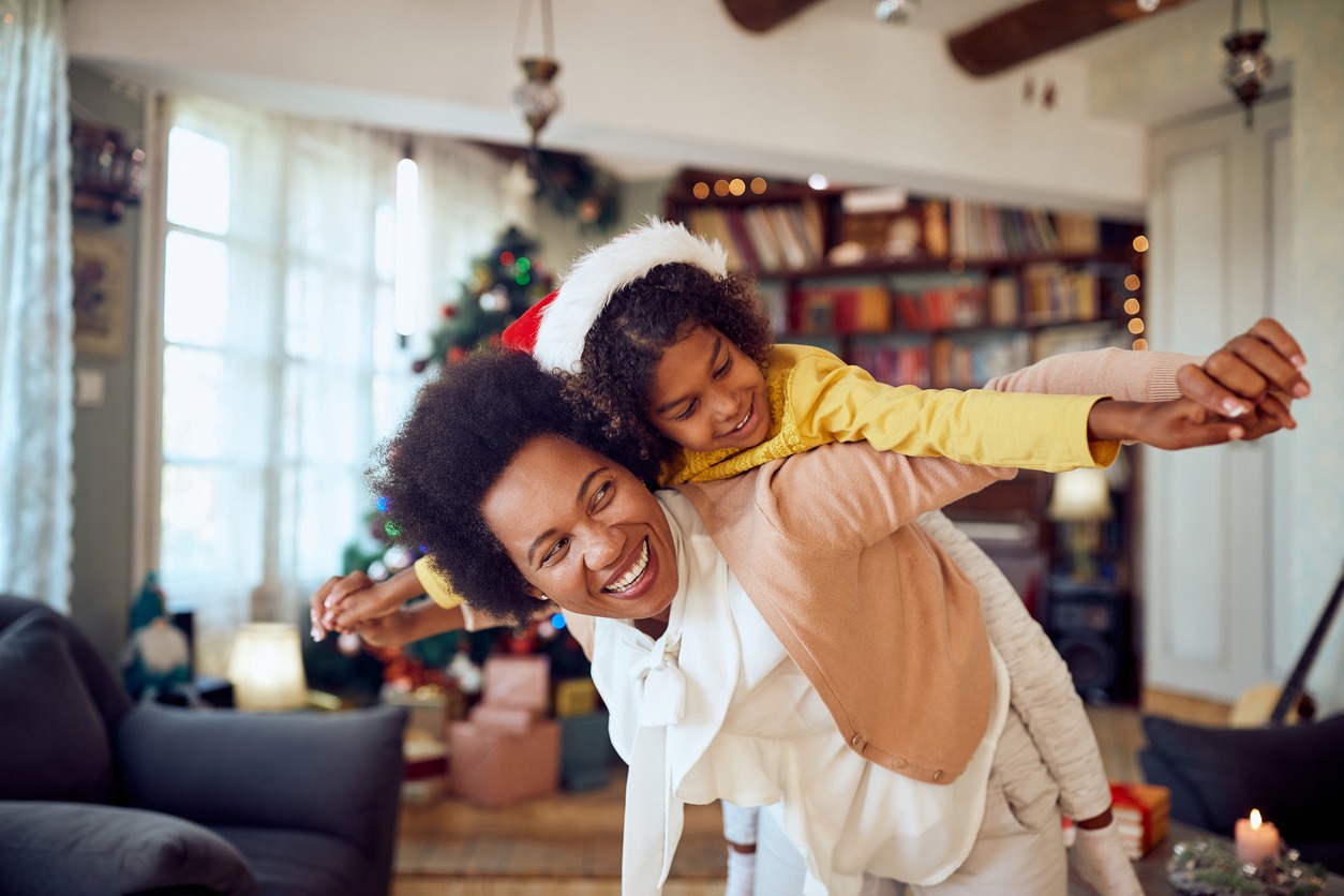 A joyful person piggybacks a child wearing a Santa hat in a festive, decorated living room with a Christmas tree in the background.