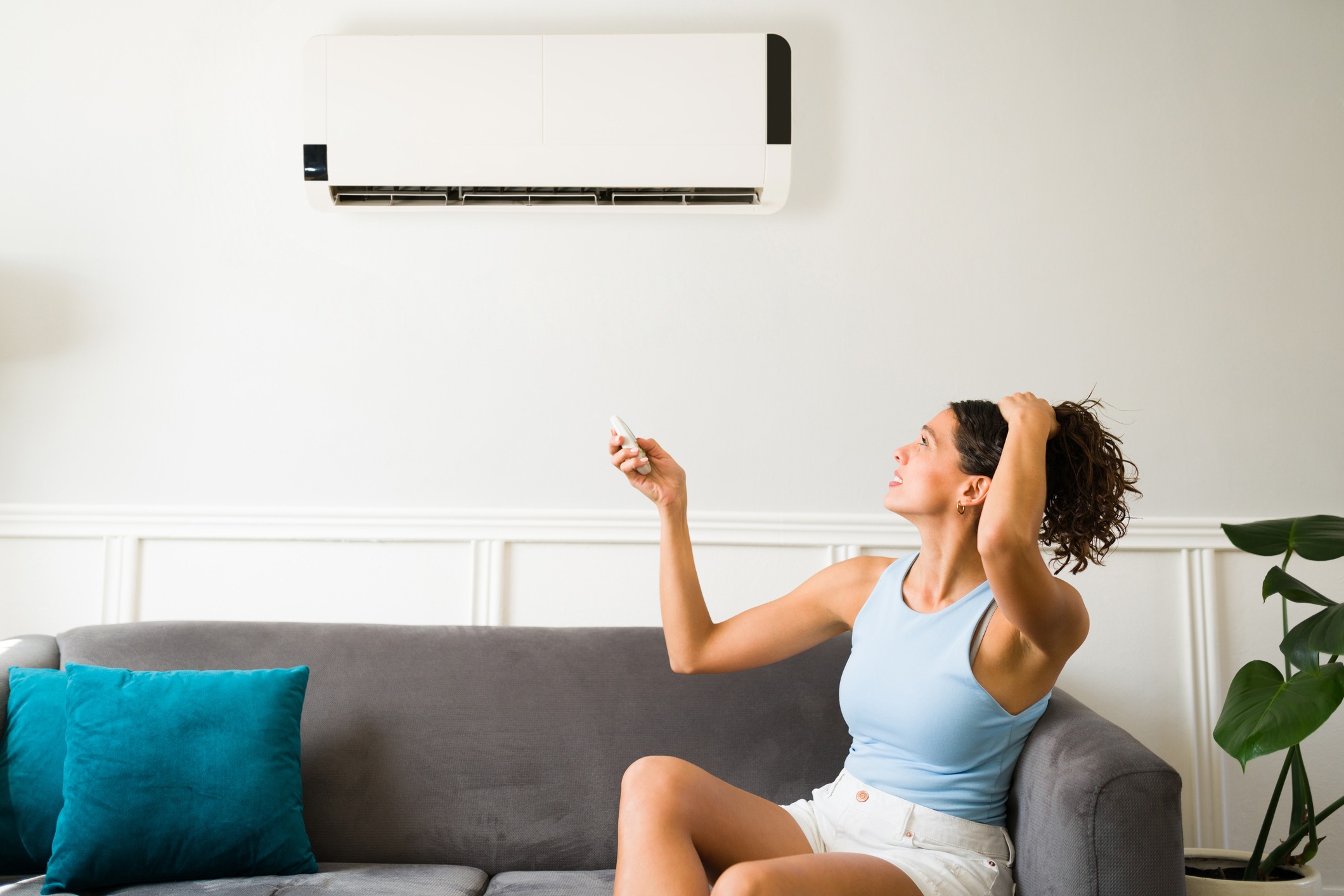A person on a sofa adjusts an air conditioner with a remote, sitting beside teal cushions and a potted plant.