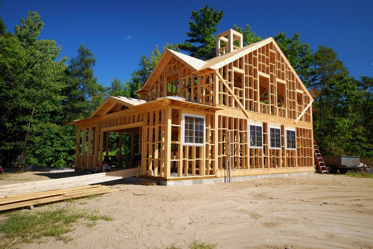 A wooden house frame stands under construction on sandy ground, surrounded by green trees, with clear blue skies above.
