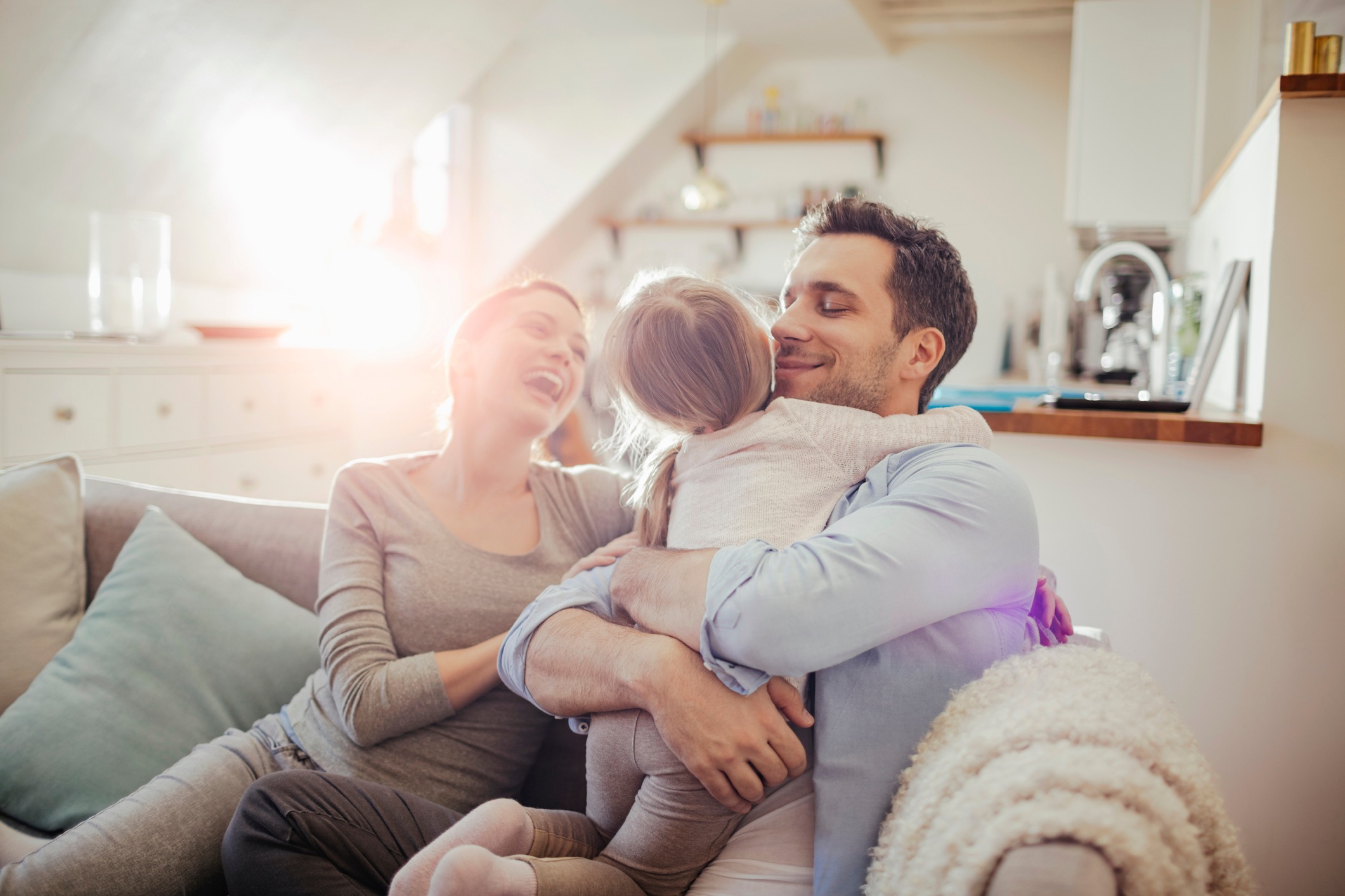 A joyful family moment featuring a person and child embracing on a cozy couch while another person laughs, bathed in warm sunlight.