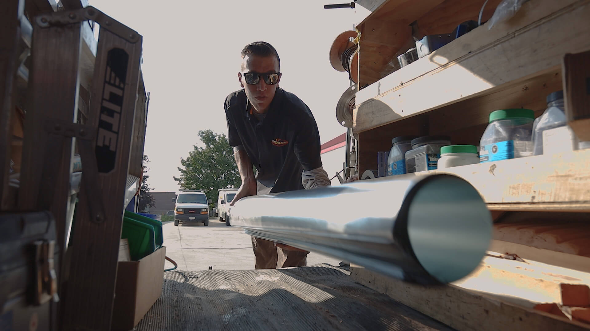 A person in sunglasses handles a large metal pipe near a vehicle, surrounded by tools and materials in a sunlit area.