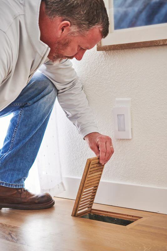 A photo of a mean kneeling on the floor, lifting up a furnace vent cover to look inside.