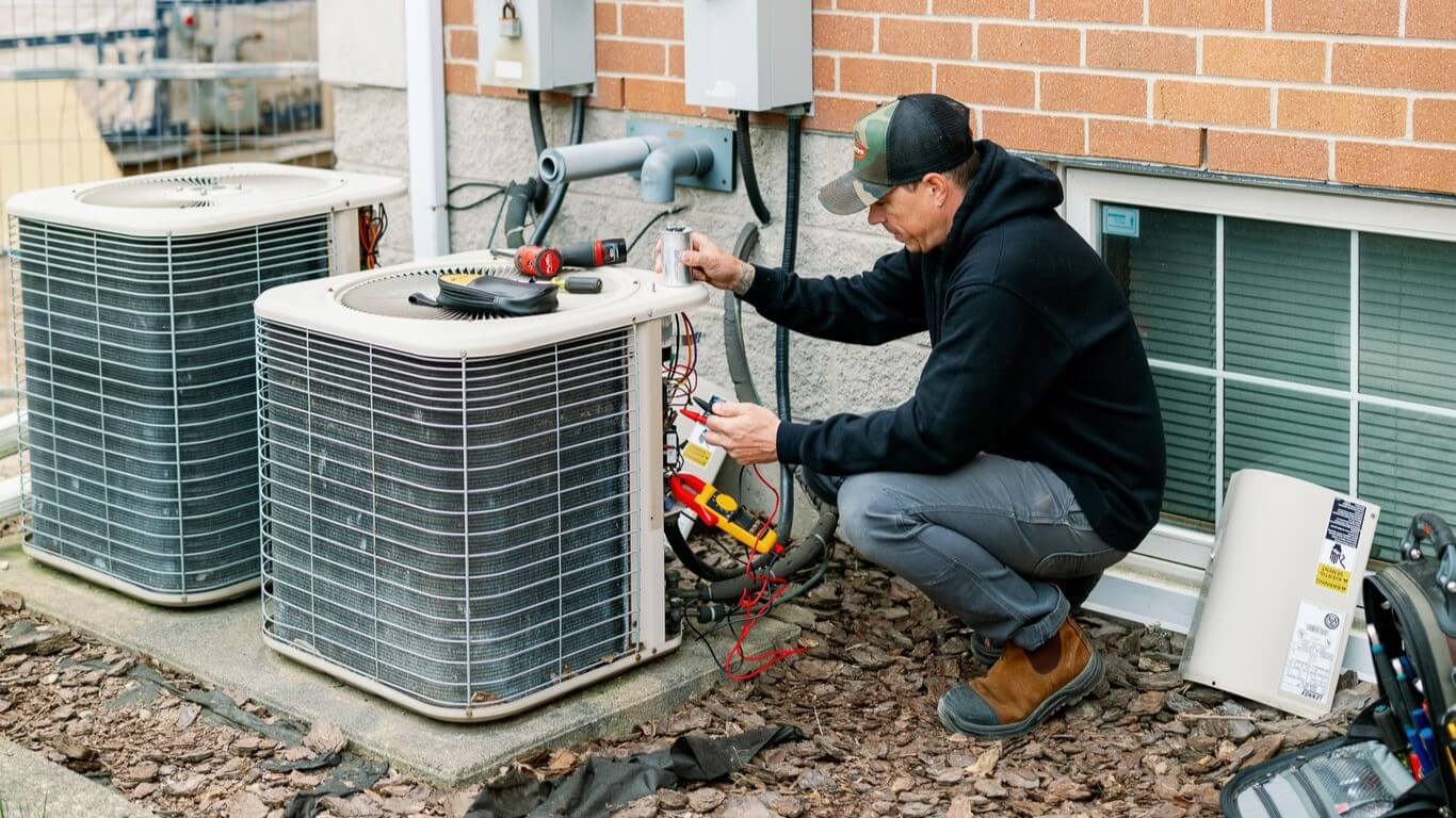 A person is repairing an outdoor air conditioning unit against a brick wall, using tools and wires. The area is covered with mulch.