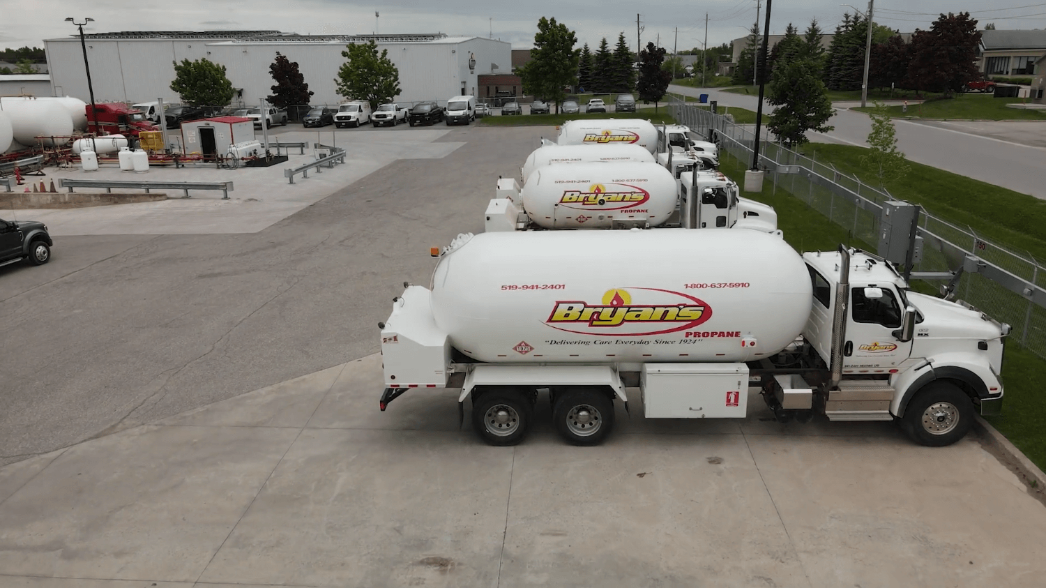 A series of parked propane tank trucks with "Bryan's" branding, adjacent to industrial buildings, trees, and parked cars in a paved area.