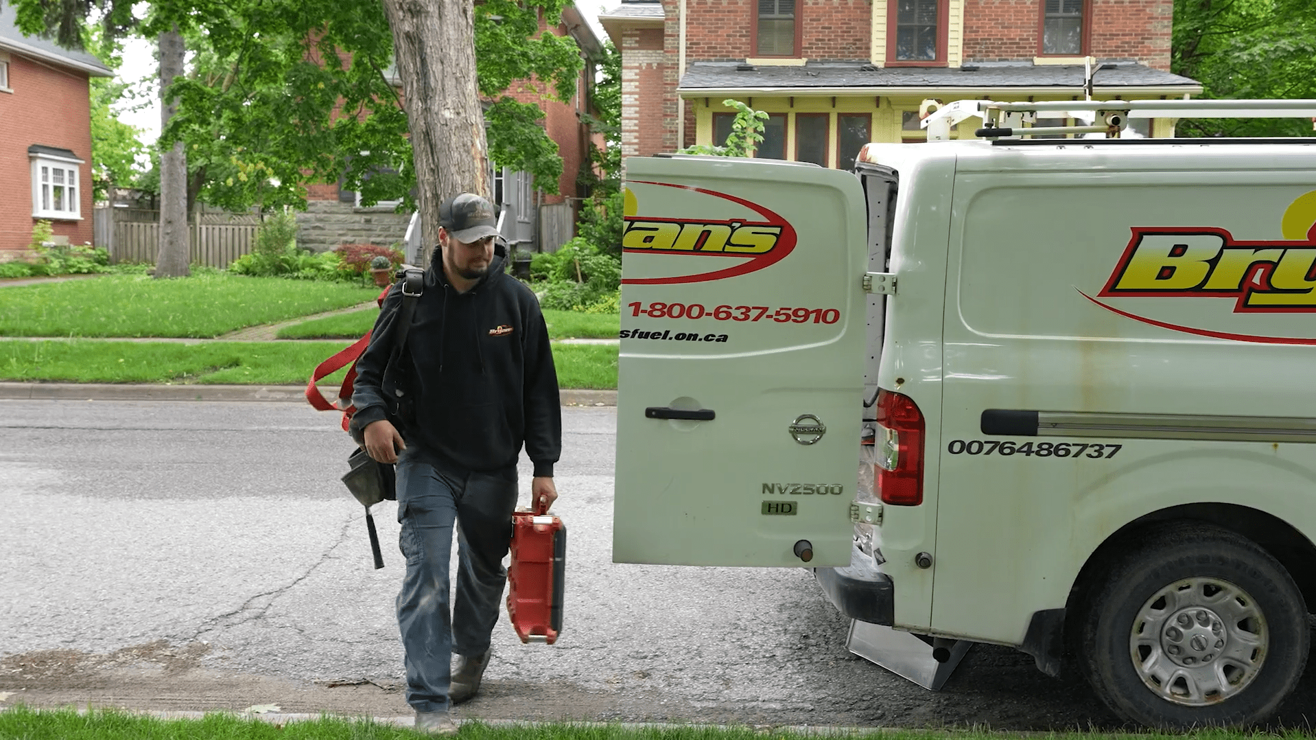 A person carrying tools walks away from a branded van parked on a residential street with brick houses and lush greenery.