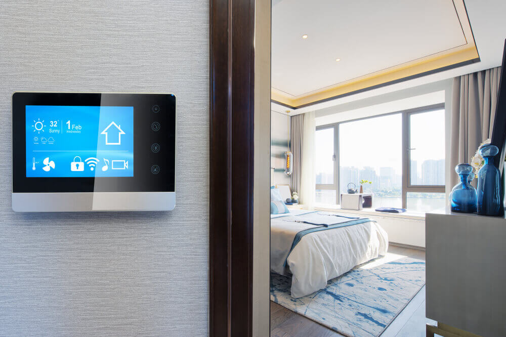 Smart home control panel in a modern bedroom with large windows, blue decor, and a comfortable bed. City skyline visible outside.