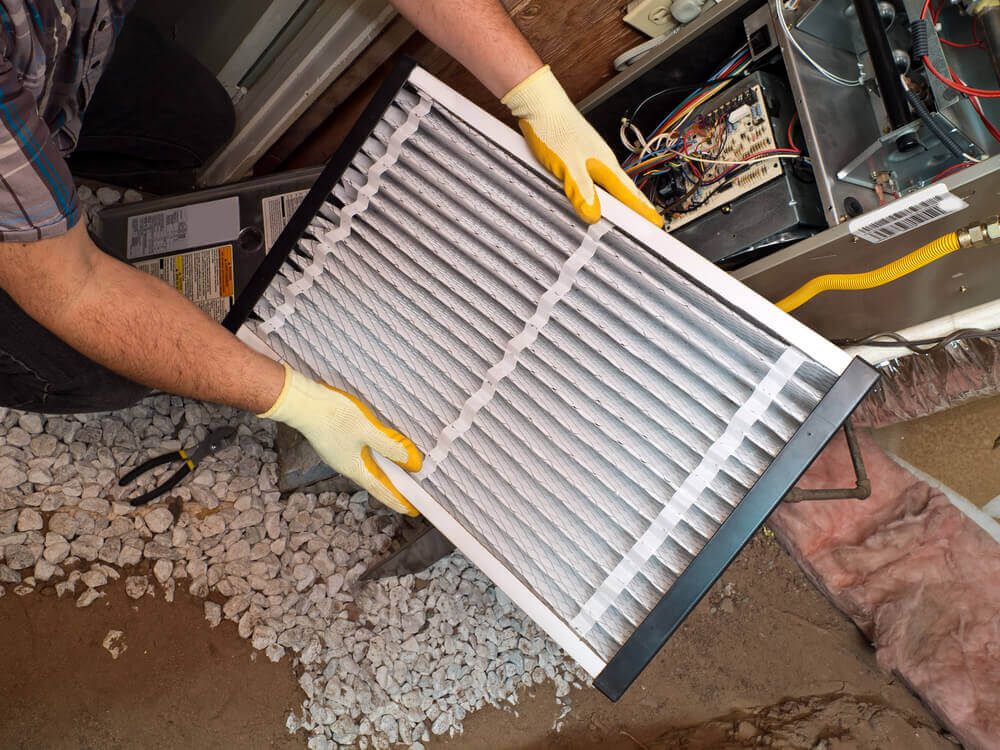 A person wearing gloves holds a large rectangular air filter near an HVAC system, surrounded by pebbled flooring and various tools.