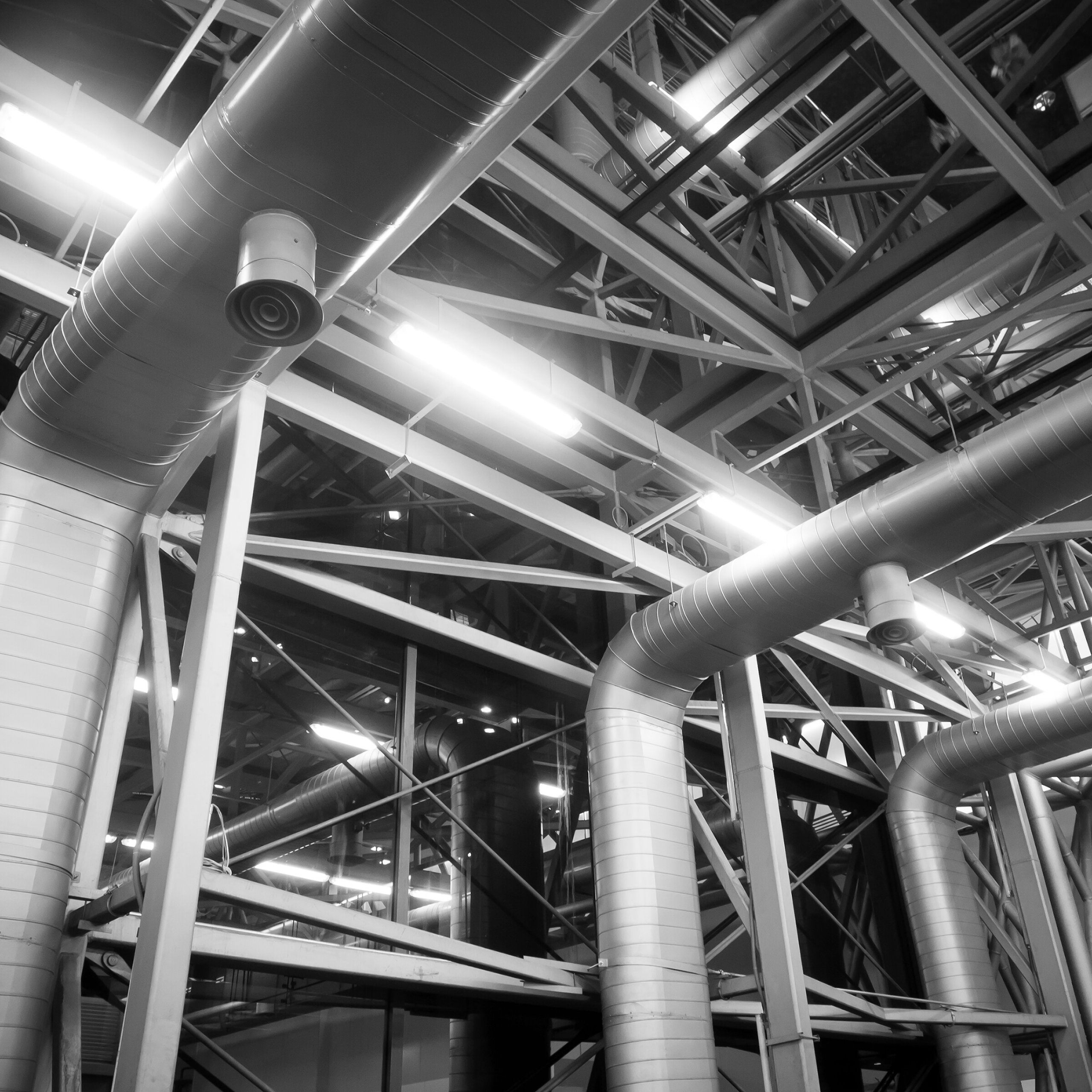 Industrial interior with large metal pipes and structural beams, featuring a high ceiling. The setting is modern and geometric, emphasizing industrial architecture.