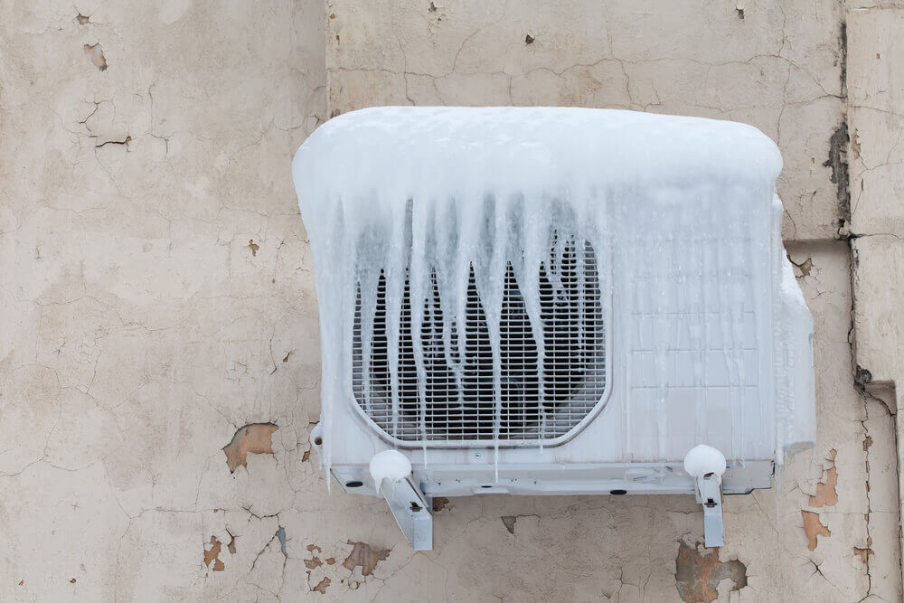 An outdoor air conditioning unit coated with ice is mounted on a peeling, weathered wall, showcasing extreme cold weather conditions.