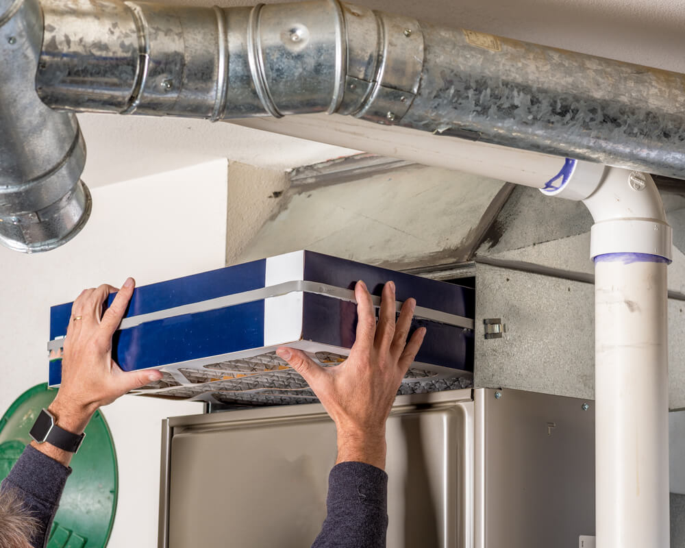 A person is installing a filter in a residential heating, ventilation, and air conditioning (HVAC) unit, with visible metal ductwork.