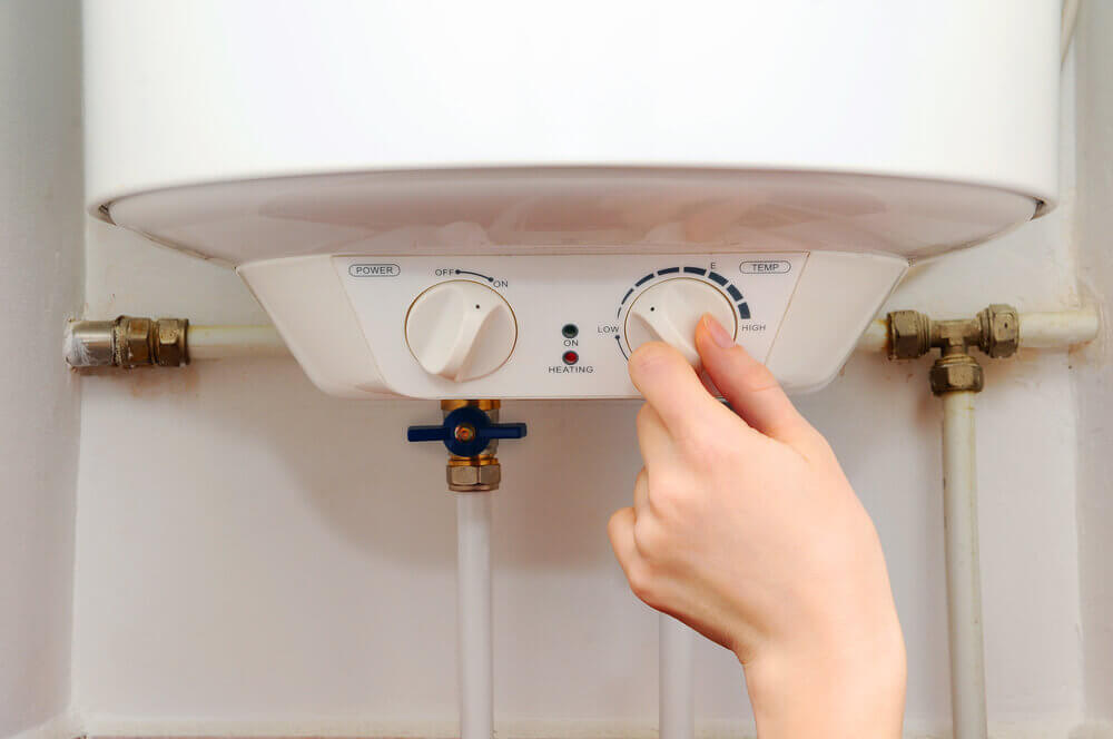 A person adjusts the temperature knob on a water heater, surrounded by pipes and fittings, setting it from low to high.