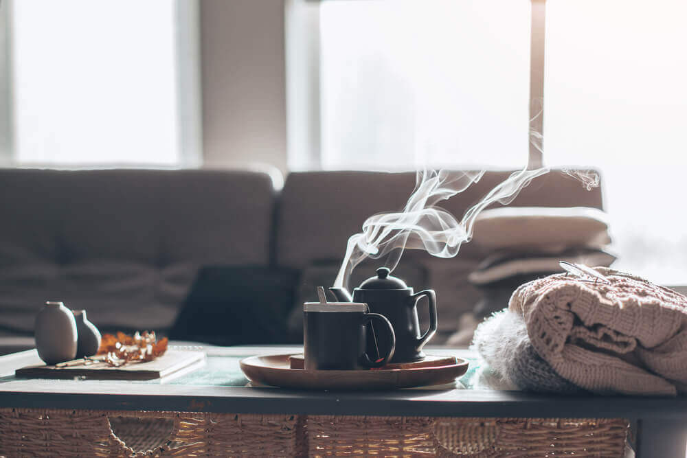A cozy living room with steaming teapot, cup, and folded blanket on a woven table, with a sofa and soft lighting.