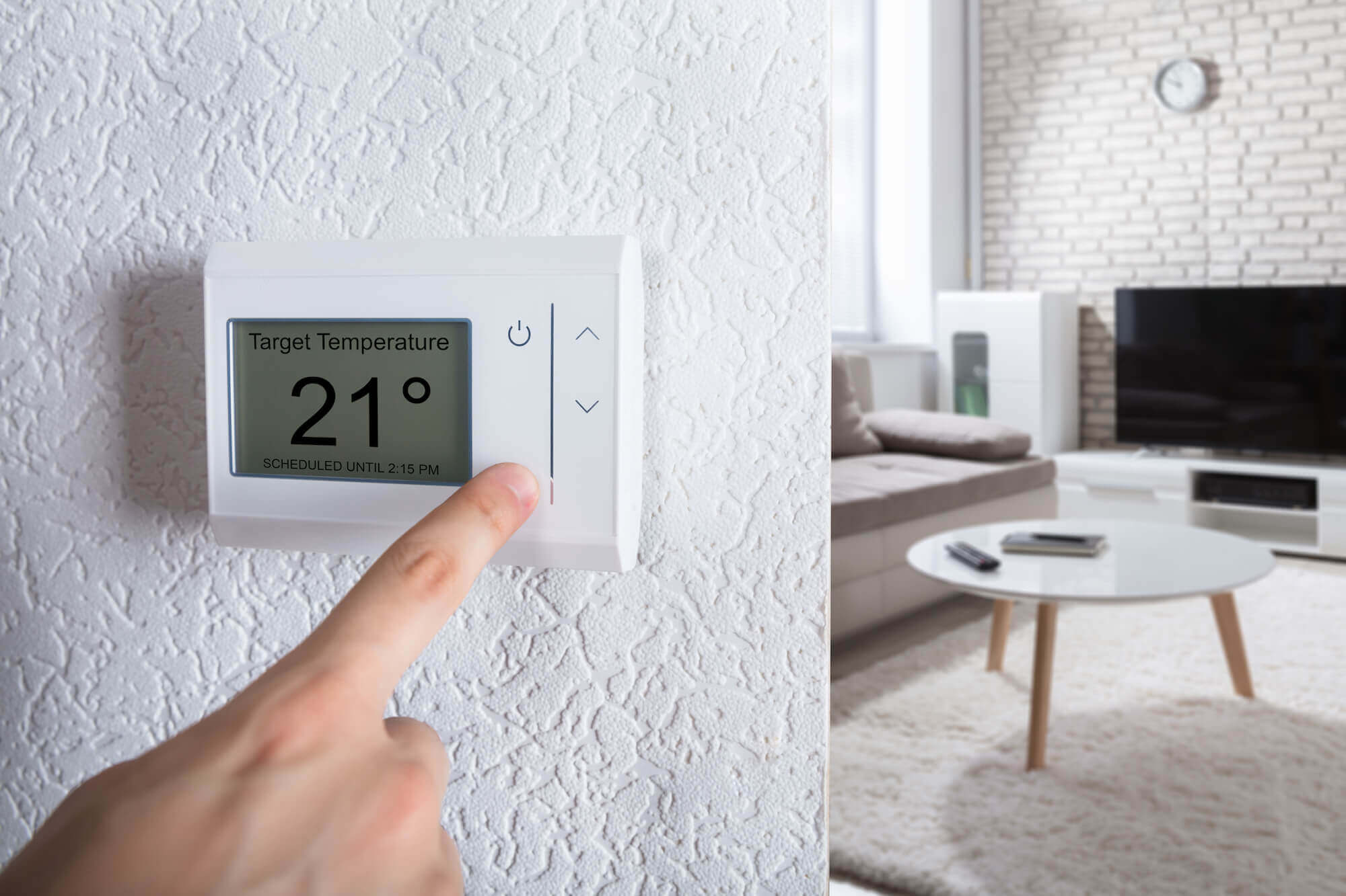 A person adjusts the temperature on a thermostat in a modern living room with a TV, table, and minimalist decor.