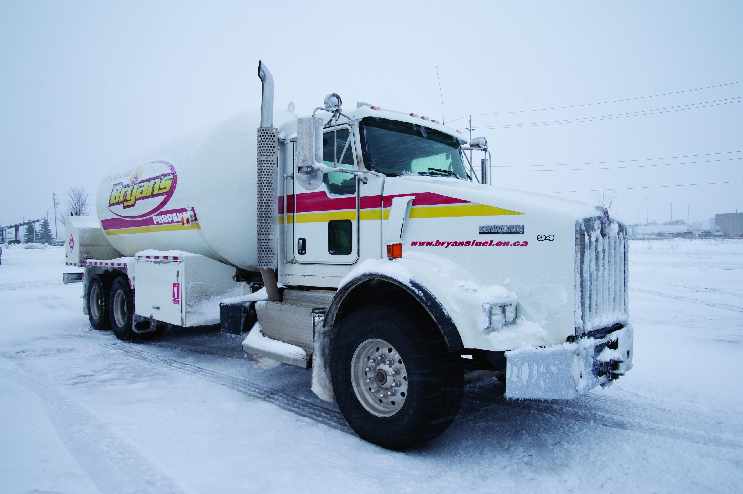 A propane delivery truck with "Bryans" branding is covered in snow, parked in a snowy, industrial area on a winter day.