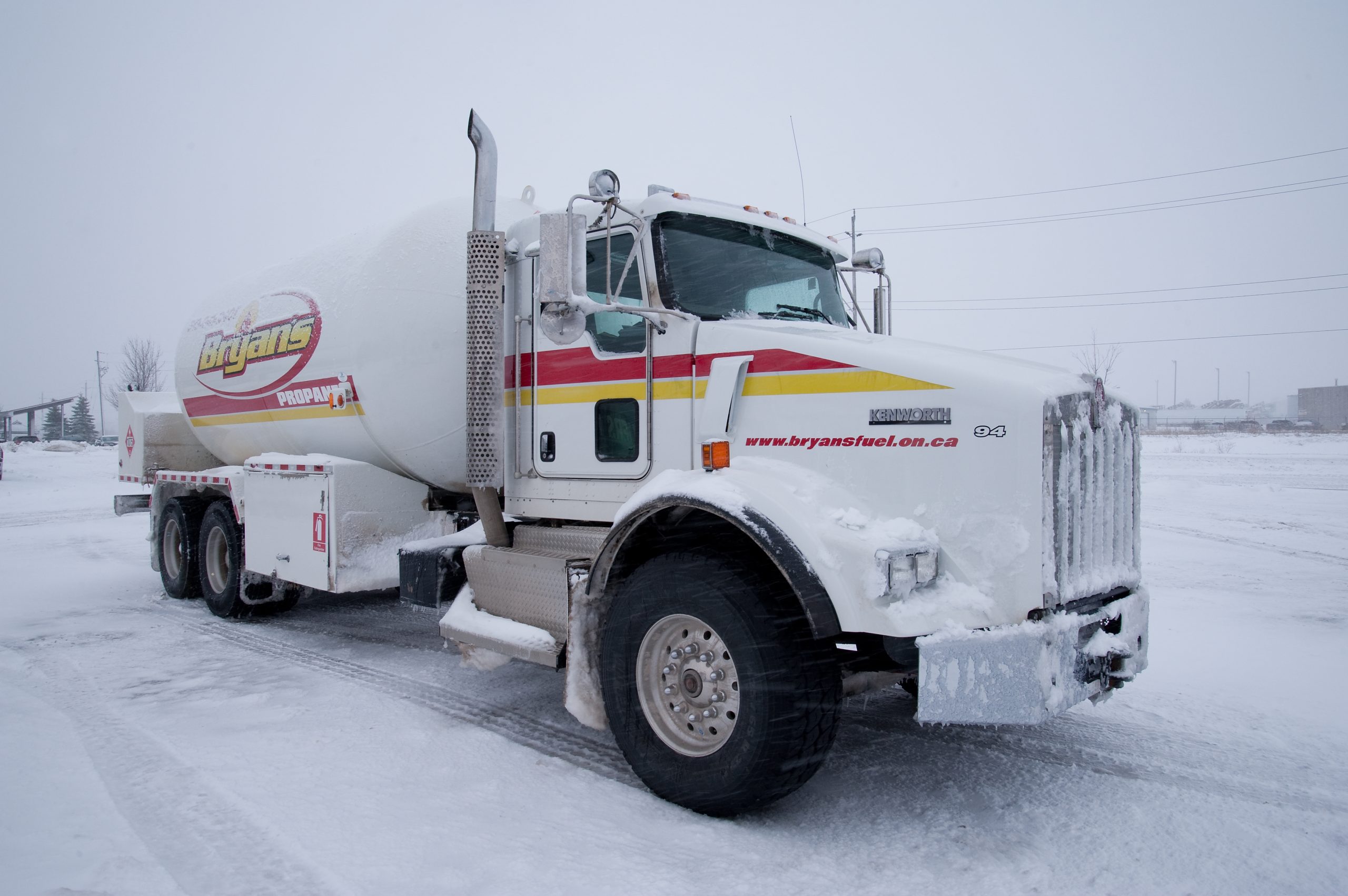 A fuel truck covered with snow is parked in a snowy area. The truck has a Bryans logo and website on the side.