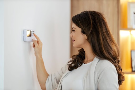 A person adjusting a thermostat on a wall in a modern, brightly lit room. The person wears a light cardigan and smiles slightly.