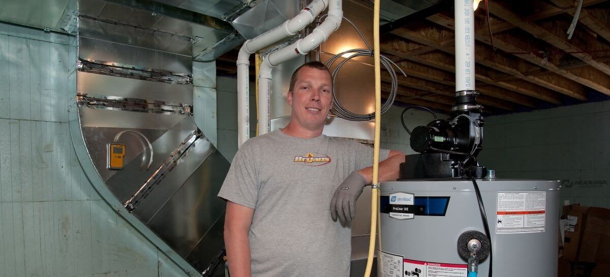 A person stands in a basement near HVAC ducts and a water heater, smiling, amid exposed beams and pipes.