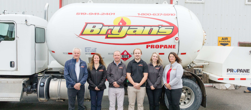 Five people are standing in front of a white propane truck with "Bryan's Propane" branding. They are wearing coordinated work attire and smiling.