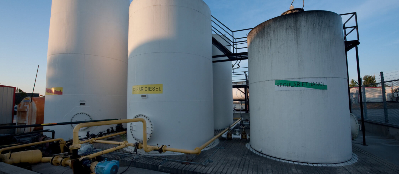 The image shows large industrial storage tanks labeled "CLEAR DIESEL" and "REGULAR ETHANOL," with pipes and pumps in the foreground during daylight.