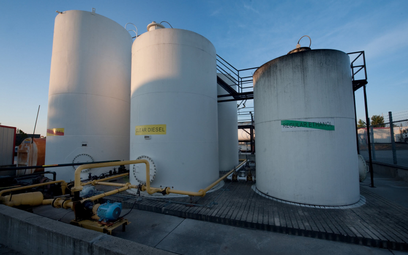 The image shows large industrial storage tanks labeled "CLEAR DIESEL" and "REGULAR ETHANOL," with pipes and pumps in the foreground during daylight.