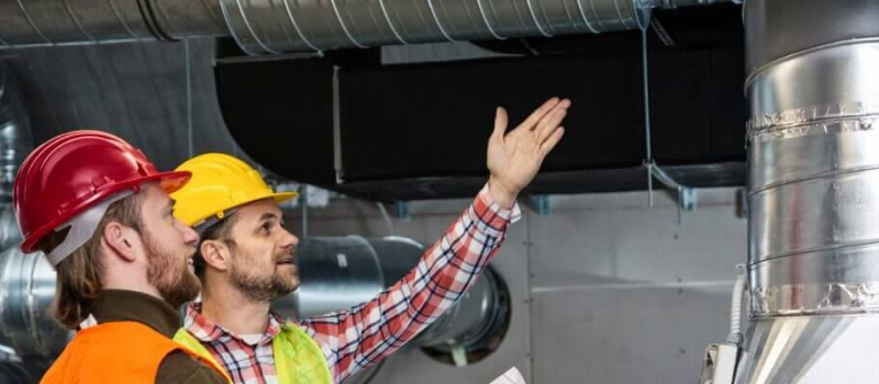 A person wearing a yellow hard hat and high visibility vest inspecting overhead ductwork, with a focused expression and gesturing upwards.