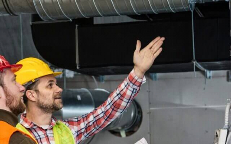 A person wearing a yellow hard hat and high visibility vest inspecting overhead ductwork, with a focused expression and gesturing upwards.