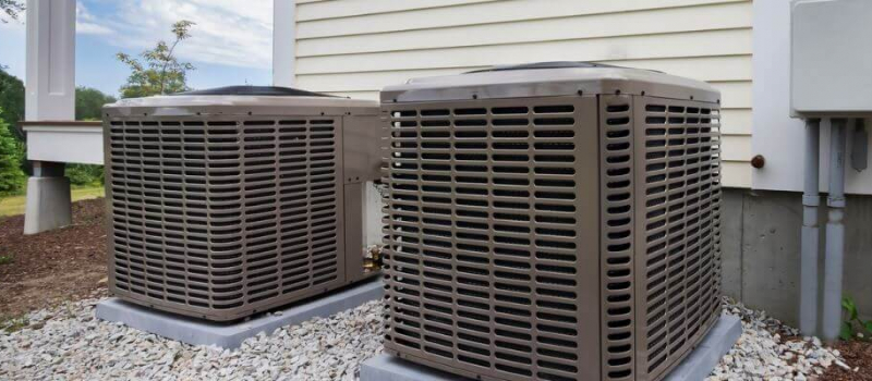 The image shows two brown outdoor air conditioning units beside a house with vinyl siding, set on a bed of white gravel.