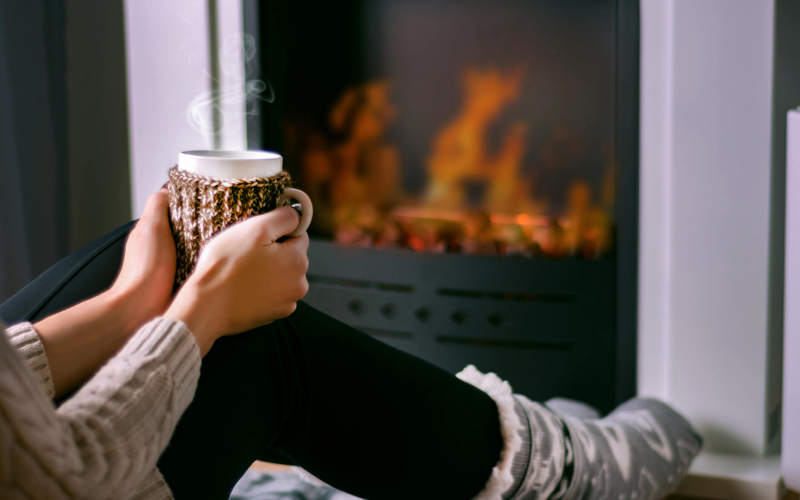 A person is holding a mug with both hands, steam rising from it, sitting comfortably by a cozy-looking electric fireplace with visible flames.