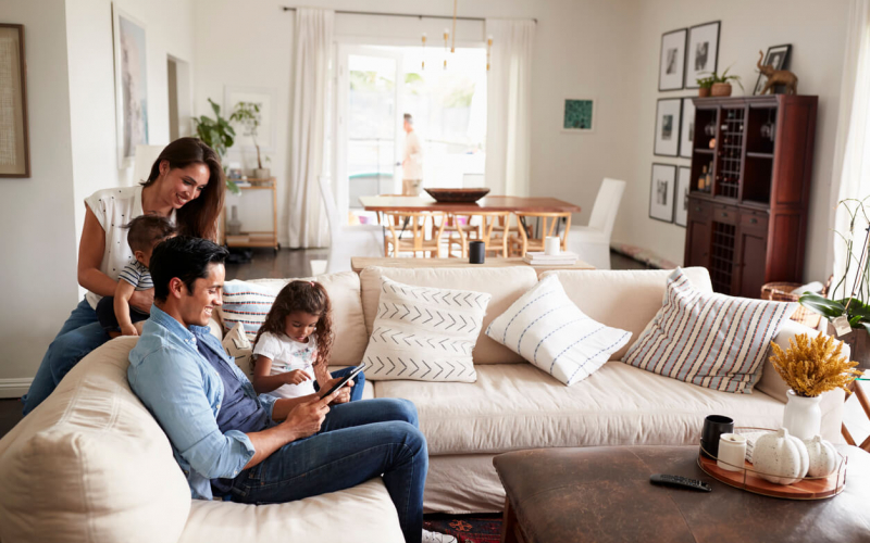 A family in a cozy living room; one person using a tablet with a child, another holding a toddler, and a third person in the background.